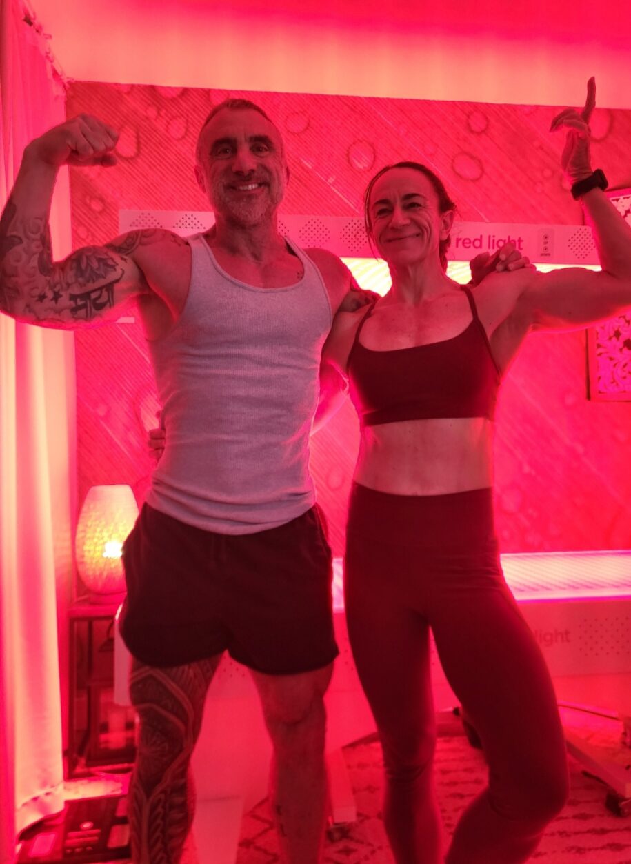Two people smiling and flexing beside an illuminated Vital Red Light Therapy bed